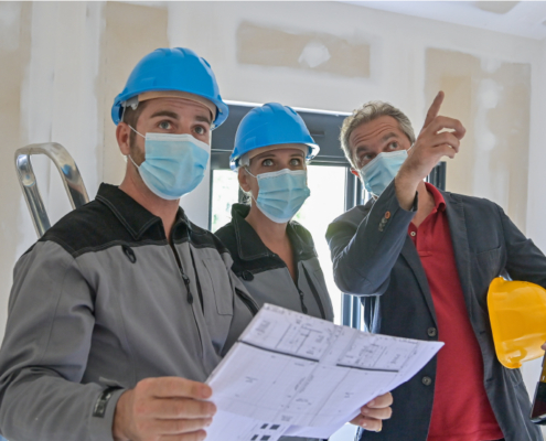 3 men in hard hats and mask, with tools, looking over blueprints